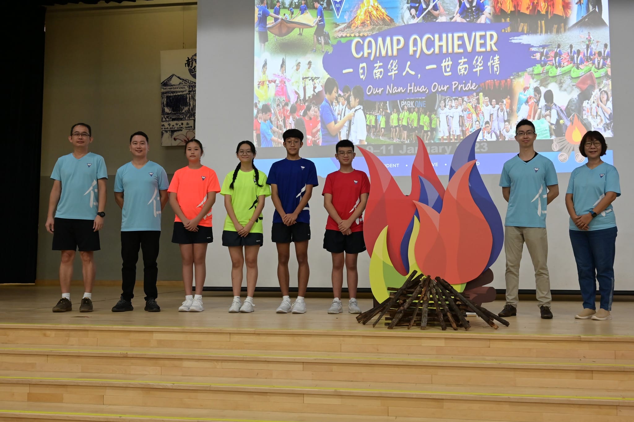 Eight people stand on a stage near a decorative campfire cutout, with a Camp Achiever event slideshow projected behind them.